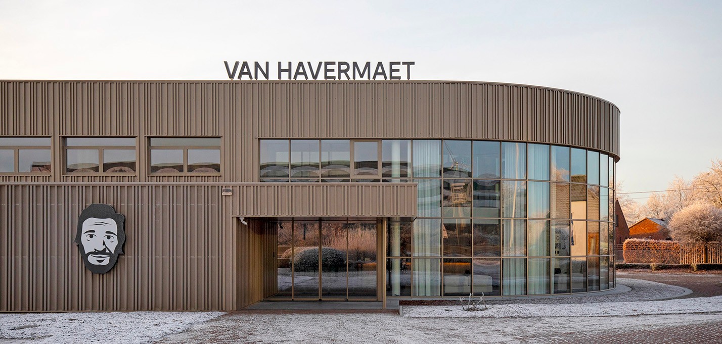Het moderne kantoor van Van Havermaet in Westerlo. Het gebouw heeft een beige/bruine kleur en grote, afrondende raampartijen op één hoek.