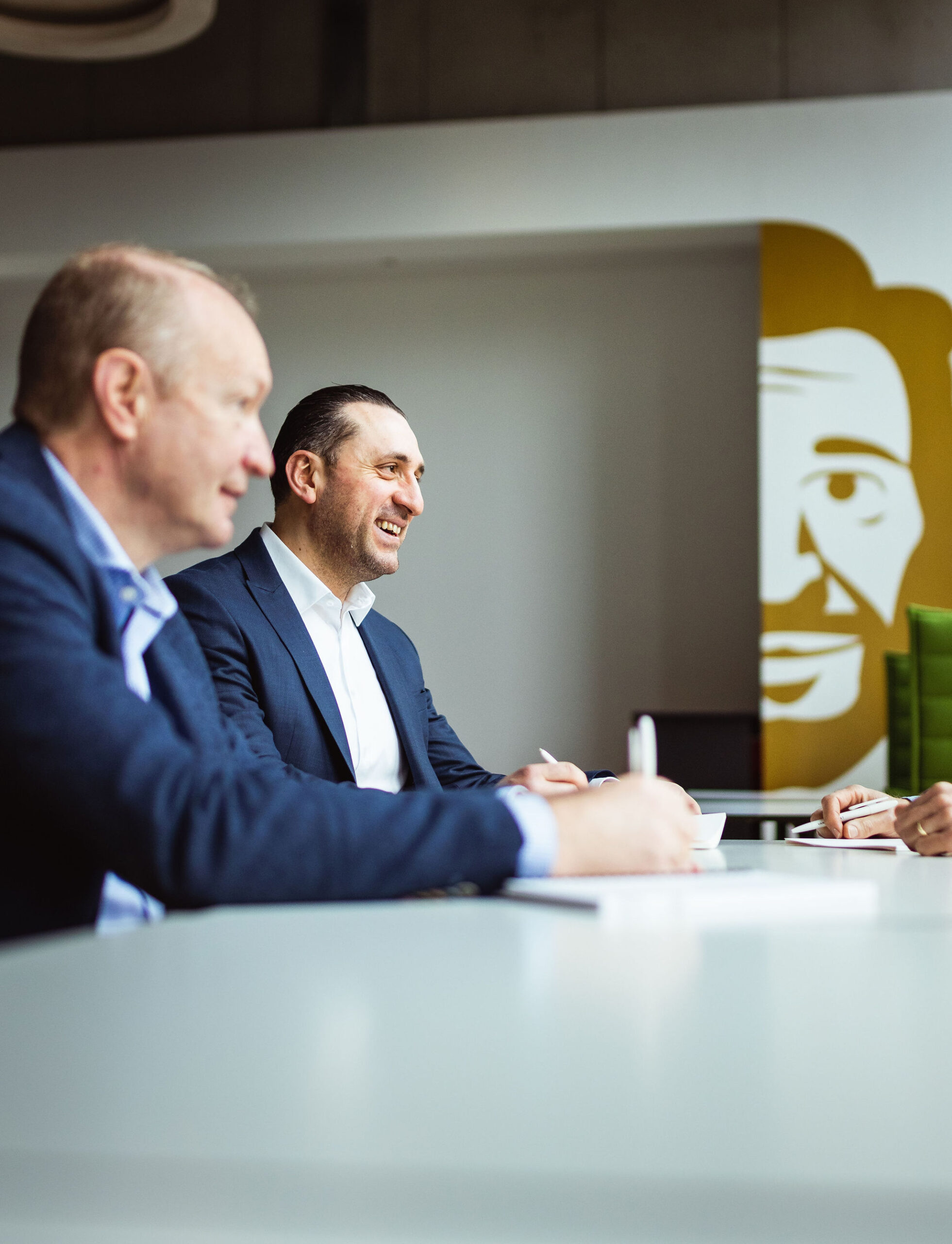 Twee collega's die werken bij Van Havermaet zitten aan tafel tijdens een vergadering. Op de achtergrond prijkt het logo van Van Havermaet in het groot.