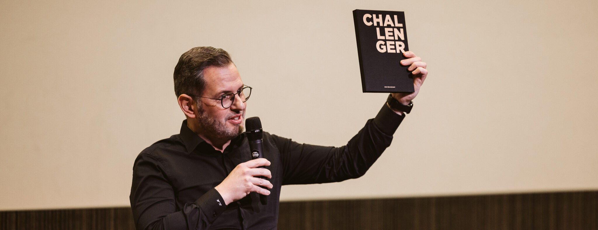 Kristof Meesters, CEO van Van Havermaet, toont tijdens een presentatie die hij geeft het boek 'Challenger'. Hij draagt een zwart hemd en bril, en spreekt in een microfoon.