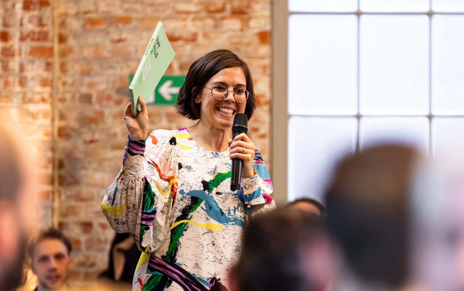 Een collega stelt tijdens een presentatie een boek voor aan haar andere collega's of maeten. Ze spreekt in een microfoon en draagt een kleurrijke blouse.